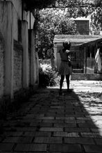 Load image into Gallery viewer, Black and white image of migrant woman walking in her courtyard in the shadows
